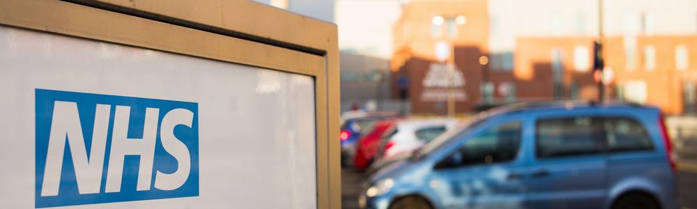 A sign says 'NHS'. A car park in the background is out of focus with a blue car visible.