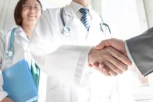 A doctor shaking hands with someone in plain clothes. Their handshake is the focus of the image. A female healthworker is smiling in the background out of focus.