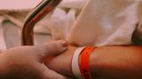 A child in hospital. Their hand is being held by an older person. The child's wrist has lots of bands on it from previous surgeries and hospital stays. The background can barely be seen, but it is a light pink.