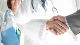 A doctor shaking hands with someone in plain clothes. Their handshake is the focus of the image. A female healthworker is smiling in the background out of focus.