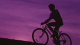 A silhouette of a person cycling up a hill against a purple sky