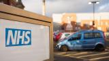 A sign says 'NHS'. A car park in the background is out of focus with a blue car visible.