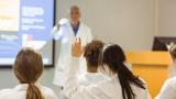 A class of medical students. One of them is raising their hands to answer a question from the lecturer.