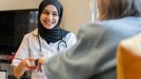A young doctor talks to a patient, smiling. She has a stethoscope around her neck.