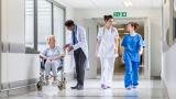 Four people in a hospital corridor. A doctor in a white coat speaking to an elderly woman in a wheelchair, and two nurses.
