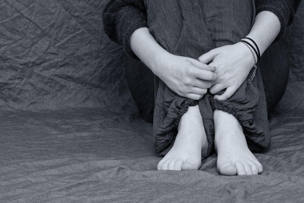 A black and white photo of a child is crouched in the corner of the room. We cannot see their face, and we only see their hands gripping their legs in a foetal position.