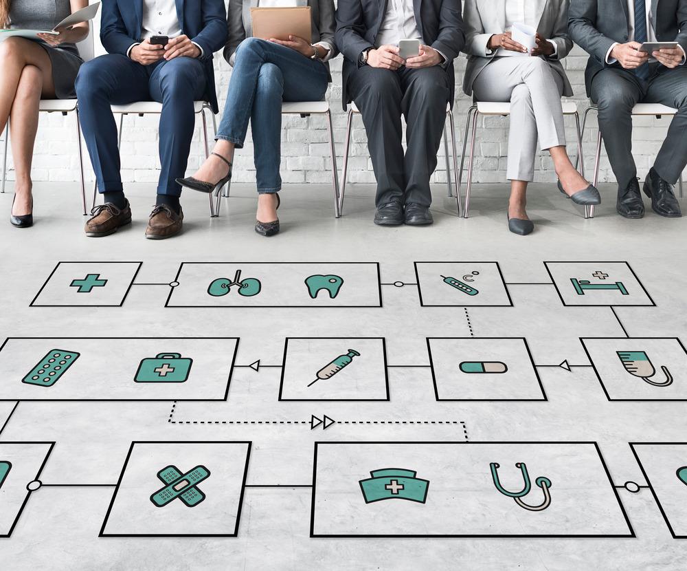 A row of people sit in a row. Only their legs can be seen. On the floor in front of them are a collection of boxes which have medical symbols inside them (eg. syringe, plaster and stethoscope).