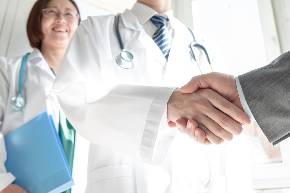 A doctor shaking hands with someone in plain clothes. Their handshake is the focus of the image. A female healthworker is smiling in the background out of focus.