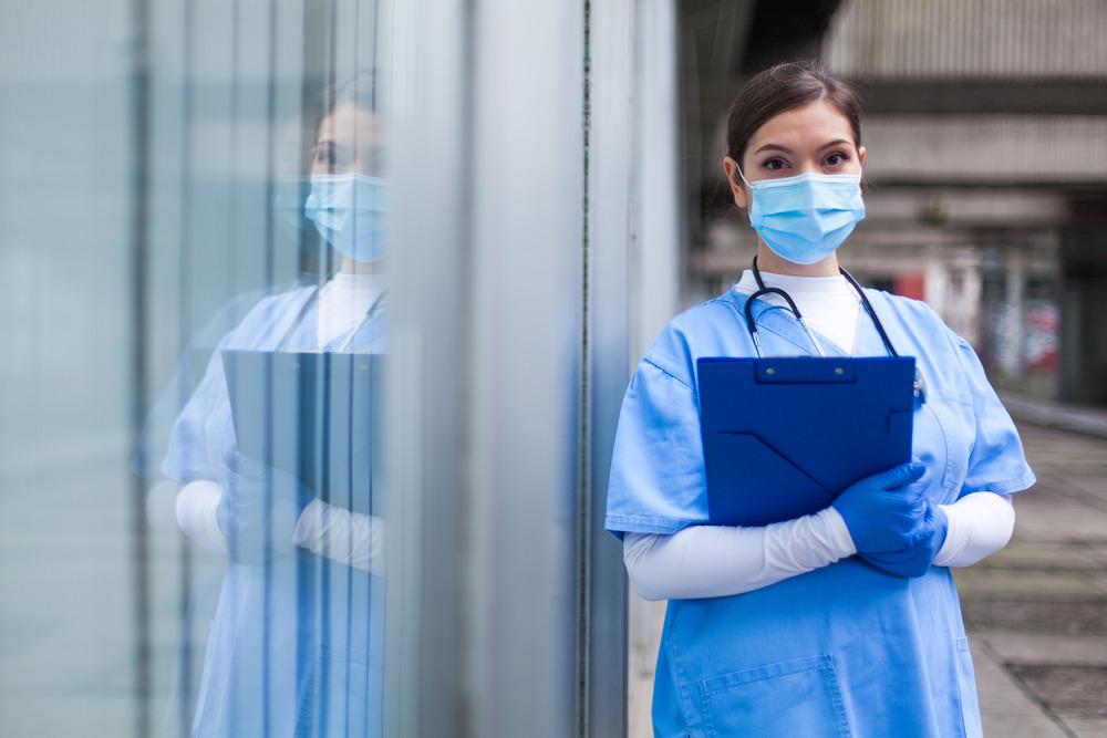 A female health worker stands next to a window, her reflection in the glass next to her. She is wearing PPE.