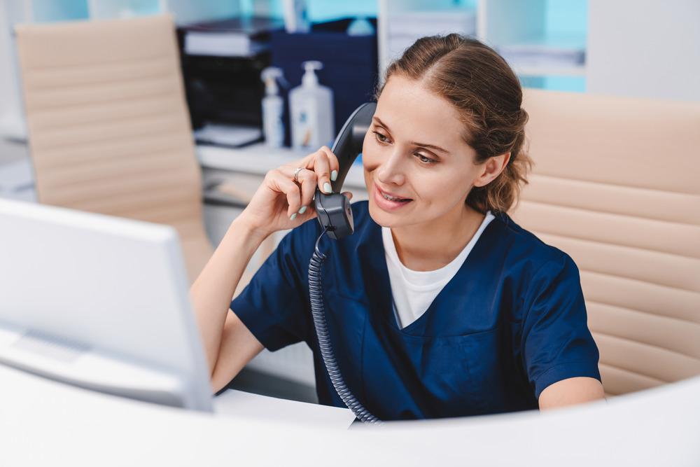 A young female receptionist is on the phone at a GP surgery. She is looking at her monitor.