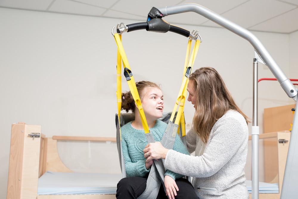 A young girl with a disability being lifted with a harness. Her carer guides her from the bed, looking at her and smiling.